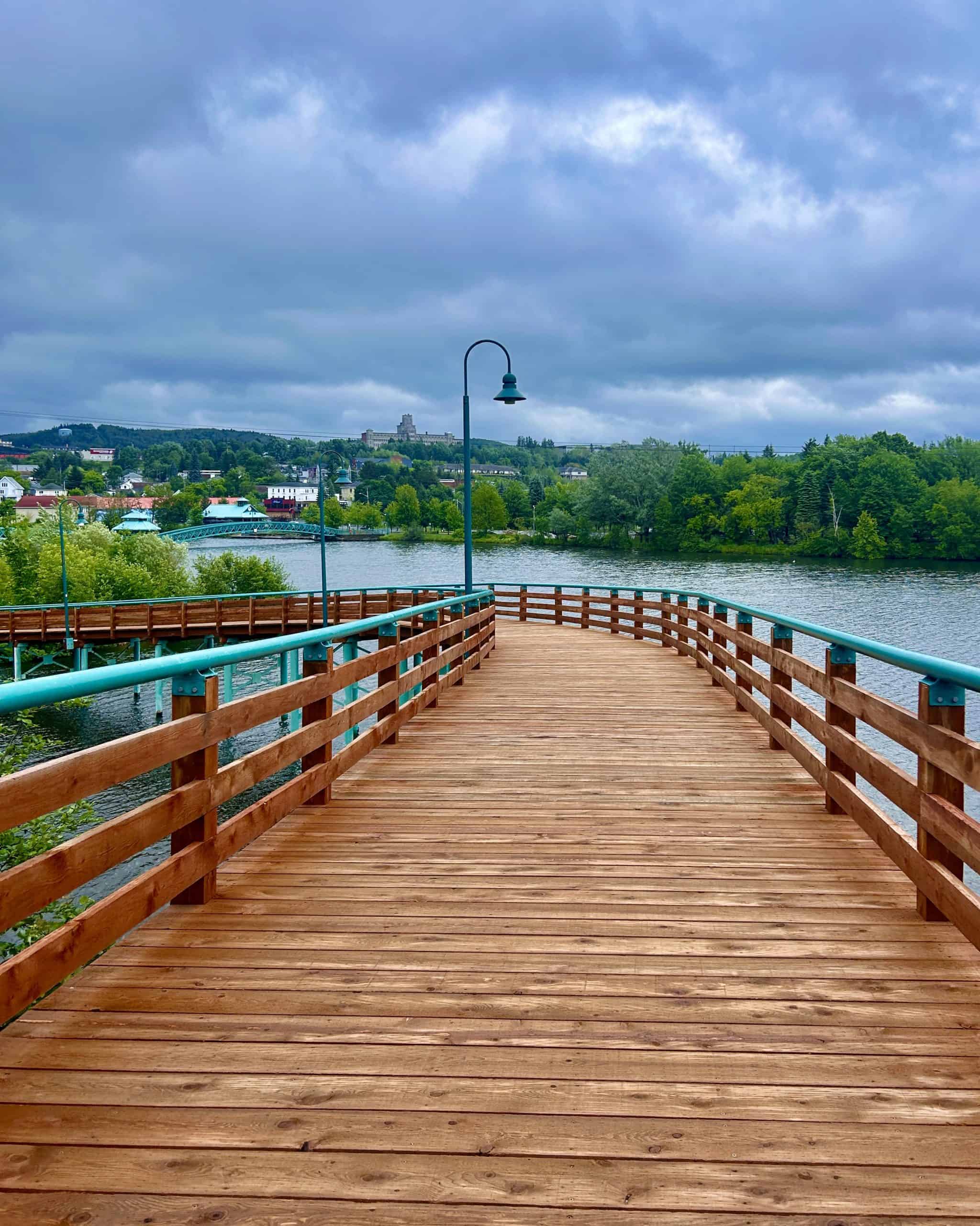 Wooden riverside boardwalk in Edmundston, New Brunswick, stretching over calm water under a cloudy sky, surrounded by green trees and small colorful houses.