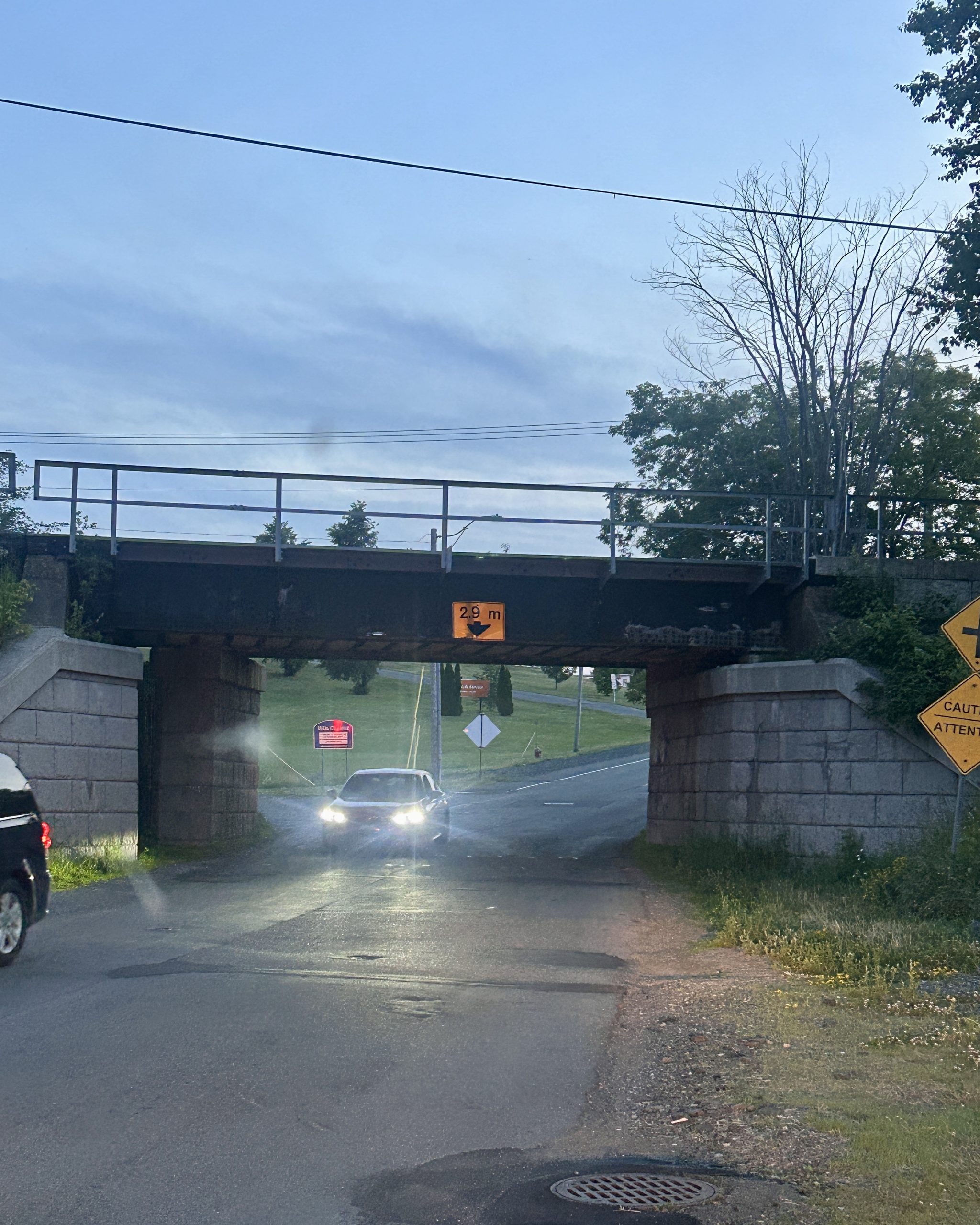 A low railway overpass with a clearance sign reading “2.9m” sits over a small road at dusk, as two cars with headlights pass underneath. The sky is slightly cloudy and trees line the background, while caution signs are visible to the right.