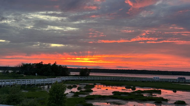 Sunset over the marshlands near Phare de l’Ile aux Foins in Neguac, New Brunswick. A wooden boardwalk stretches across calm waters, with vibrant orange, pink, and purple skies reflecting on the surface under a dramatic cloudscape.