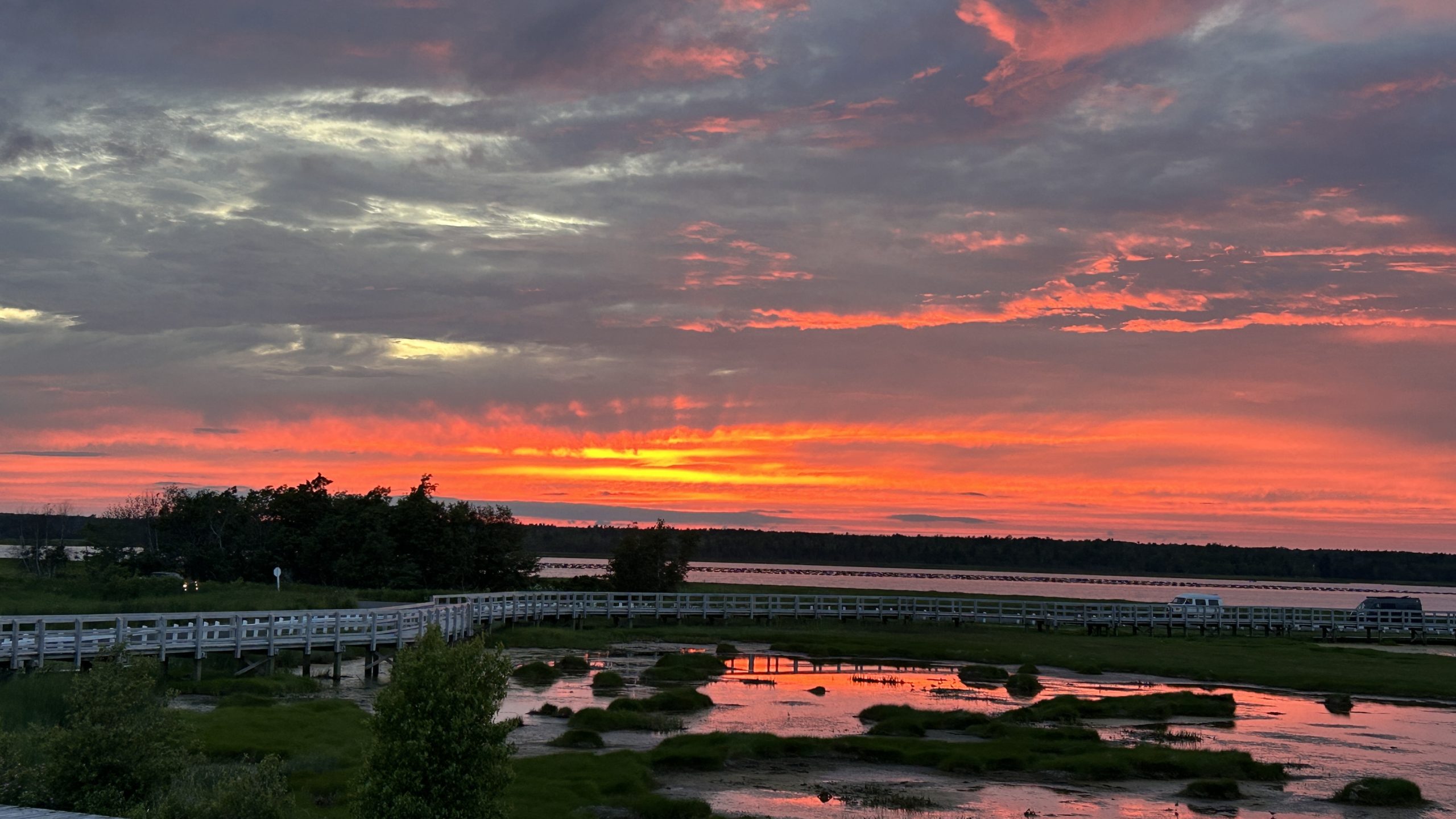 Sunset over the marshlands near Phare de l’Ile aux Foins in Neguac, New Brunswick. A wooden boardwalk stretches across calm waters, with vibrant orange, pink, and purple skies reflecting on the surface under a dramatic cloudscape.