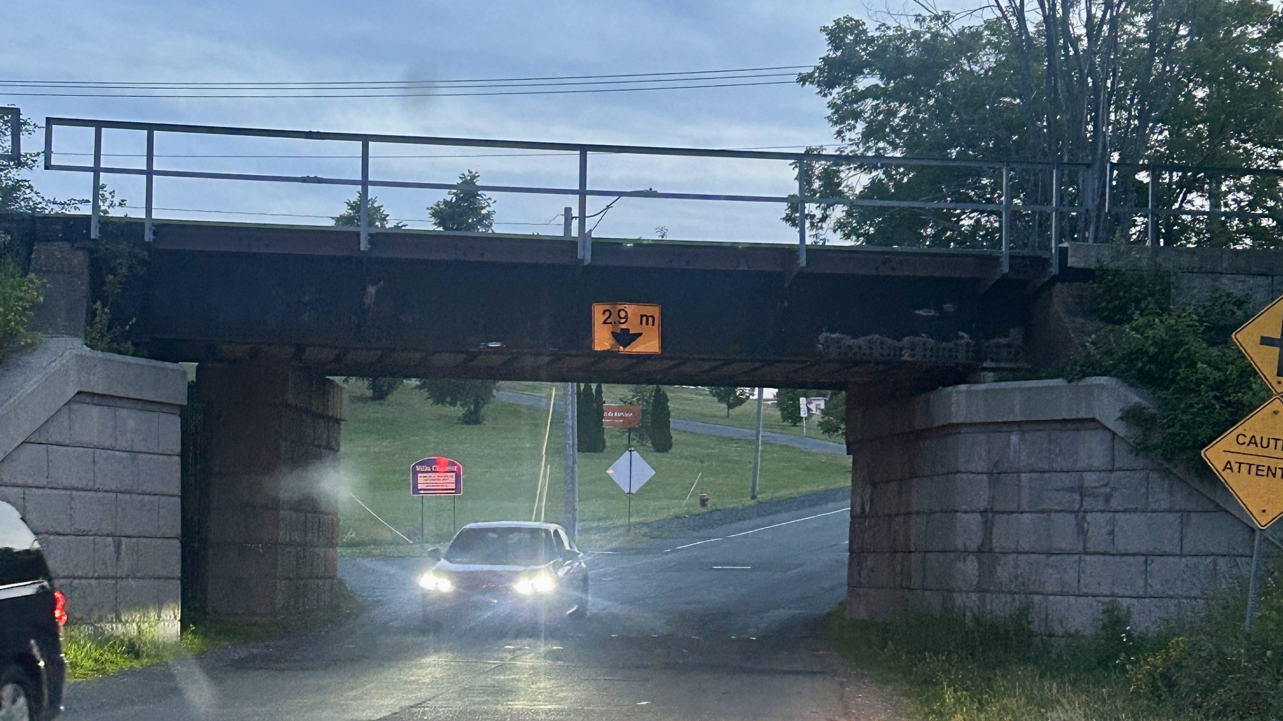 A low railway overpass with a clearance sign reading “2.9m” sits over a small road at dusk, as two cars with headlights pass underneath. The sky is slightly cloudy and trees line the background, while caution signs are visible to the right.
